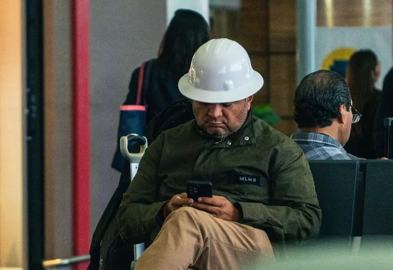 Trabajador con casco blanco, sentado, mira un video en su celular