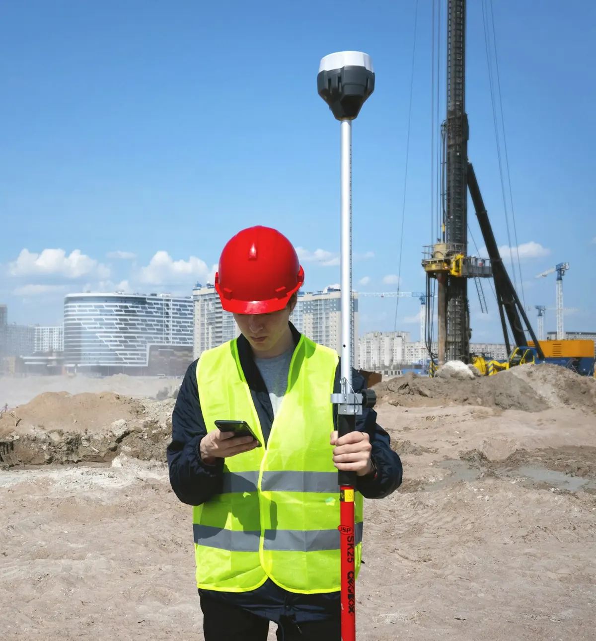 Trabajador con casco rojo y chaleco reflectante mira su celular en una obra a pleno sol